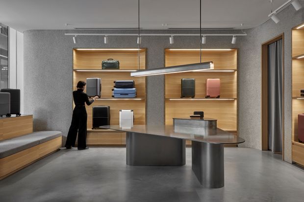 Contemporary showroom interior displaying luggage and travel cases on illuminated timber shelving. The space features polished concrete flooring, grey textured walls, integrated display joinery, a dark sculptural central display table, track lighting, and a built-in bench along the left wall.