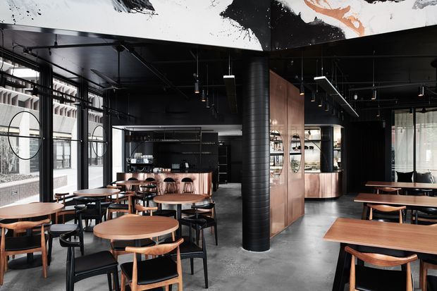 A stylish dark-toned commercial dining and hospitality interior with numerous timber tables and black-framed chairs arranged across an open-plan space. Black ceilings, exposed services, polished concrete flooring, black metal-framed glazing, a central cylindrical structural column, and warm timber cabinetry create a refined industrial atmosphere. Abstract black-and-white artwork spans the ceiling bulkhead, while a bar or service counter is visible at the rear.