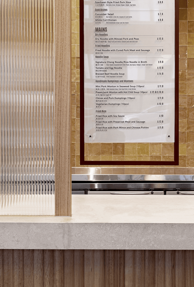 Close-up architectural detail of a commercial food-service interior, showing a framed bilingual noodle restaurant menu mounted on small ochre-brown tiles, beside a vertically ribbed translucent glass partition with timber framing. Below are stainless-steel service or counter elements, a pale stone/concrete ledge, and vertical timber wall panelling.