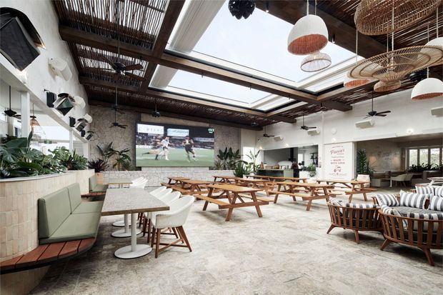 A spacious hospitality-style dining and lounge interior with numerous timber picnic tables, upholstered bench seating, armchairs, and striped lounge furniture. The room features a large glazed roof/skylight structure, exposed dark timber overhead screening, woven pendant lights, ceiling fans, abundant indoor plants, a stone-textured floor, and a wall-mounted sports screen.