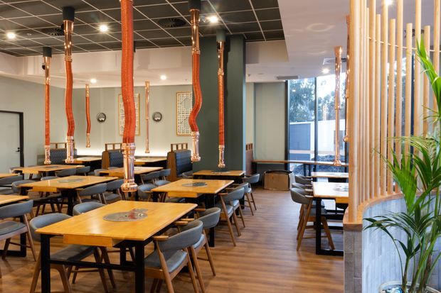 A warmly lit contemporary restaurant or café dining area with numerous timber tables and upholstered chairs arranged beneath distinctive suspended copper-toned pendant lights. The space features wood-look flooring, a dark grid ceiling, pale walls, large windows, and a vertical timber slat screen with indoor planting separating the dining zone from the glazed edge.