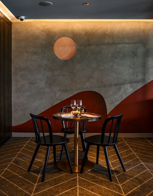 A compact, moody dining nook with a small round table set for two, black spindle-back chairs, and a brass-toned pedestal base. The space features a dramatic textured grey plaster wall with sweeping rust-red painted forms, dark timber wall panelling, warm concealed perimeter lighting, and patterned dark carpet flooring. The overall atmosphere is intimate and refined, with a strong Art Deco-inspired interior character.