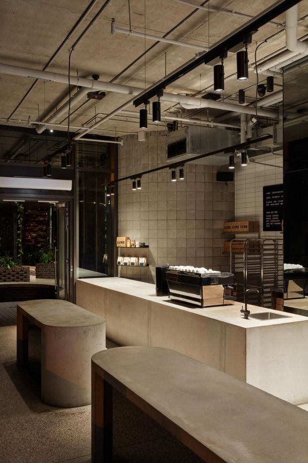 A moody industrial-style commercial café interior with a concrete service counter, rounded concrete benches, tiled walls, exposed ceiling services and ducts, black track-style pendant lighting, stainless-steel café equipment, and large glazed openings. The space has a refined, minimalist fit-out with warm low lighting and dark metal detailing.