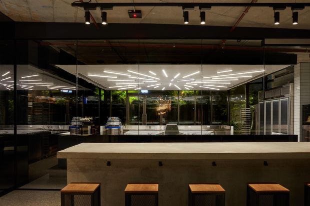A dark, contemporary commercial bar or food-service interior with a long concrete counter and timber-topped stools in the foreground. Behind the counter are stainless-steel appliances, glass-front refrigeration, white tiled walls, extensive mirrored or glazed surfaces, and dramatic linear LED ceiling lighting arranged in radiating patterns. Exposed services and dark ceiling elements create an industrial atmosphere, while greenery is visible beyond the glazing.