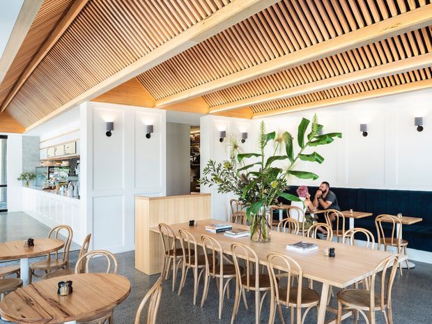 Bright contemporary hospitality-style dining space with long light-wood communal tables, rounded wooden chairs, a built-in dark teal banquette, indoor tropical planting, and a white paneled service counter. The ceiling is a prominent architectural feature, formed by exposed timber beams and closely spaced timber slats, creating a warm linear canopy over the open dining area.