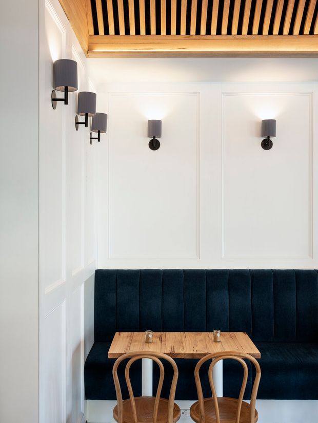 A compact, refined dining nook with a deep navy upholstered banquette, small rectangular timber dining table, and two bentwood chairs. White walls feature decorative rectangular panel mouldings and evenly spaced dark wall sconces. Above, a warm timber slatted ceiling with black recessed gaps creates a strong architectural feature. The space is softly and evenly lit, with a high-contrast palette of white, dark navy, black, and natural timber.
