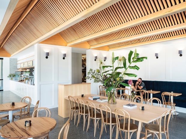 A bright, spacious hospitality-style dining and café interior with long timber dining tables, curved timber chairs, a dark upholstered banquette, indoor plants, white paneled walls, and a dramatic timber slatted ceiling with exposed beams. A service counter and shelving are visible at the rear, while large glazing brings in natural light. The palette is warm timber, white, green, and navy, creating a refined yet inviting atmosphere.