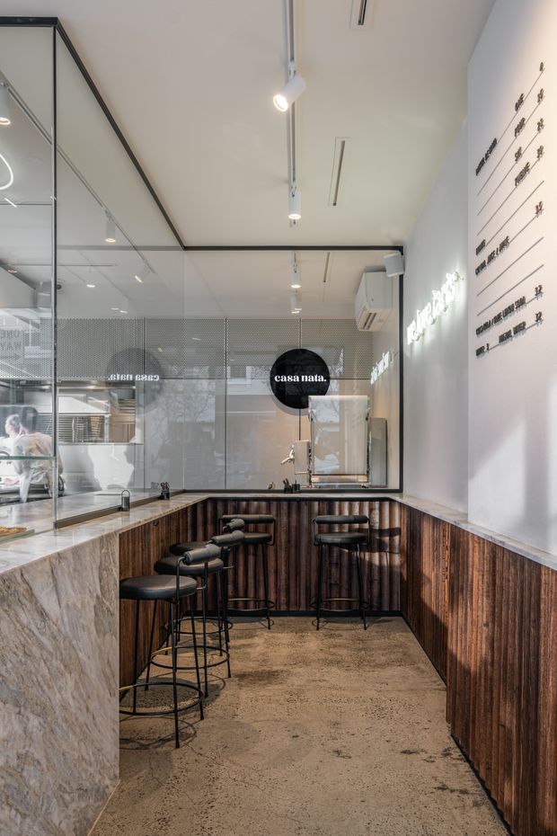 A compact contemporary commercial café or hospitality interior with a narrow bar seating area. The space features dark timber wall panelling and bar fronts, pale stone benchtops, black metal-framed glass partitions, polished concrete flooring, white ceilings, linear track lighting, and black bar stools. A menu-style wall graphic and café branding are visible, with kitchen/service areas behind the glazing.