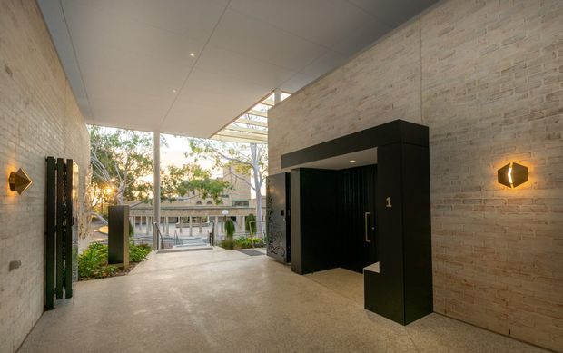 A covered contemporary entry passage with pale exposed brick walls, a smooth concrete floor, and a large black architectural doorway surround. Warm geometric wall sconces illuminate the corridor, while a glazed and open end reveals landscaped outdoor space and a multi-level building beyond. The design combines robust masonry with crisp dark joinery and a partially translucent roof structure.