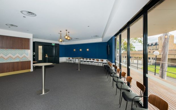 A spacious contemporary commercial lounge or hospitality area with a deep navy feature wall, patterned timber and geometric wall paneling, dark carpet, high standing tables, a long counter with stools, and rows of black-framed seating beside full-height glazing. The glazing opens visually to a timber deck and landscaped outdoor area. Decorative pendant lights and exposed ceiling services add subtle character.