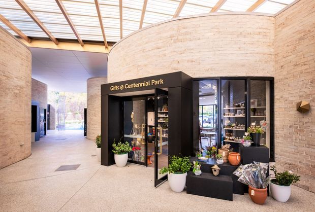 A bright contemporary commercial retail or visitor-centre interior with curved pale brick walls, a translucent roof supported by timber beams, and a black built-in gift shop kiosk labelled “Gifts @ Centennial Park.” Display shelving, potted plants, flowers and merchandise surround the kiosk, while a broad polished concrete circulation space leads toward a glazed outdoor opening.