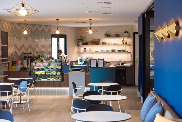 A contemporary café interior viewed from the seating area, with a service counter, illuminated refrigerated display case, coffee equipment, open shelving, and small round tables with chairs. The space features a geometric chevron-patterned feature wall, turquoise counter fronts, pale timber-look flooring, blue accent walls, and warm geometric pendant lighting. A glazed entrance or storefront is visible on the right.