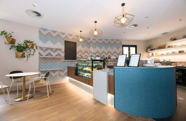 Bright contemporary café interior with a compact dining area, curved blue service counter, refrigerated display cabinet, coffee equipment, open shelving, and a bold multicoloured geometric chevron feature wall. Warm timber-look flooring, white walls and ceiling, hanging geometric pendant lights, and wall-mounted planter boxes create a lively yet clean fit-out.