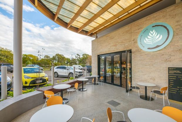 Exterior covered terrace of a contemporary café or commercial venue, with outdoor café tables and orange chairs, a glass entrance, exposed timber canopy structure with translucent roofing, and a light brick wall bearing café signage. The terrace overlooks a car park and landscaped surroundings.