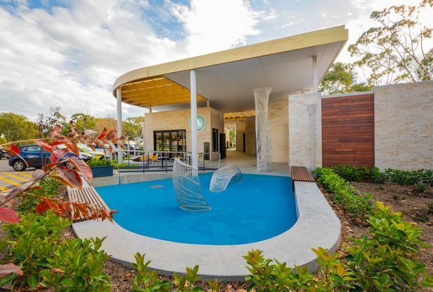 A contemporary outdoor pool courtyard beside a modern pavilion. The bright blue curved pool is surrounded by a pale concrete edge, tropical planting, timber screening, and sculptural wire chairs. The pavilion features a broad flat roof with a curved canopy section, slender white columns, exposed warm timber soffit detailing, and large glazed openings.