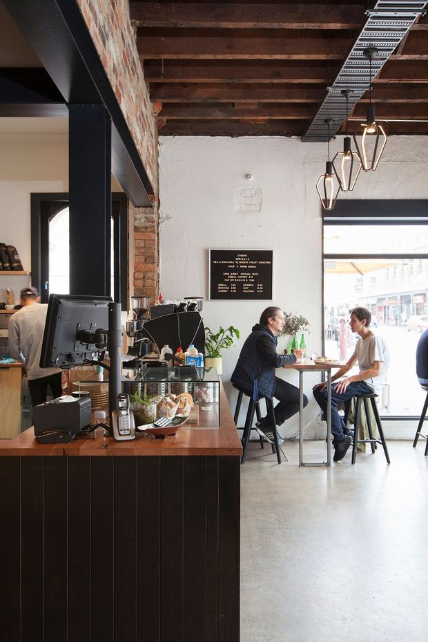 Interior view of an urban café with a dark timber service counter and coffee equipment in the foreground, communal seating by a large front window, exposed brick and white textured plaster walls, black steel framing, exposed timber ceiling beams, and hanging geometric pendant lights. Customers are seated at a small table, with a streetscape visible through the glazing.