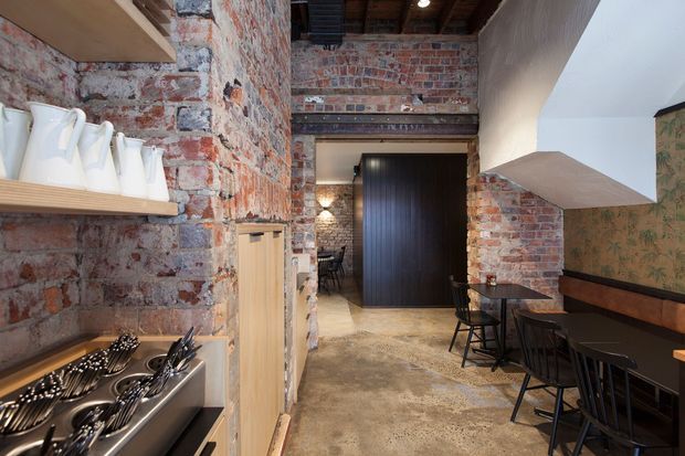 A narrow industrial-style commercial dining space, likely a café or restaurant, with exposed red brick walls, dark timber ceiling beams, concrete flooring, timber open shelving, and a long built-in bench with black tables and chairs. A serving or utensil station occupies the foreground, while a dark timber doorway and additional seating extend toward the rear. A large angular ceiling bulkhead or canopy defines the right side of the space.