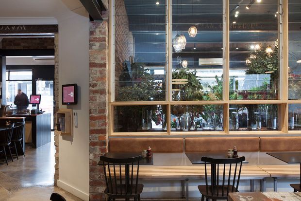 A stylish urban restaurant or café interior with a built-in banquette dining area in the foreground. The space features exposed red brick, timber-framed internal glazing, dark exposed ceiling structure, pendant lights, indoor planting, and black spindle-back dining chairs. A bar or service area is visible through the opening on the left.