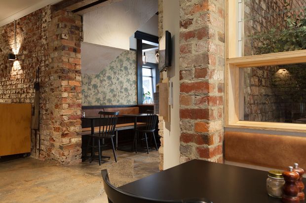 Warm industrial-style interior with exposed red brick walls, rough concrete flooring, and dark timber or metal furnishings. The foreground contains a dark dining table with a tan upholstered bench, while a secondary dining nook with black chairs sits beside a botanical-patterned wall. Timber-framed internal glazing, exposed structural elements, and wall-mounted lighting contribute to the converted-workshop character.