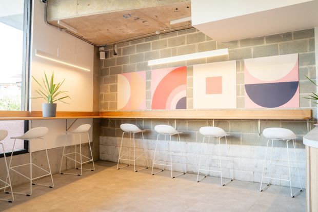 Bright contemporary communal workspace or hospitality interior with a long timber counter and white bar stools set against an exposed concrete block wall. Geometric pastel artwork, linear wall lighting, large glazing, a timber ceiling panel, and a potted plant create a playful, modern atmosphere.