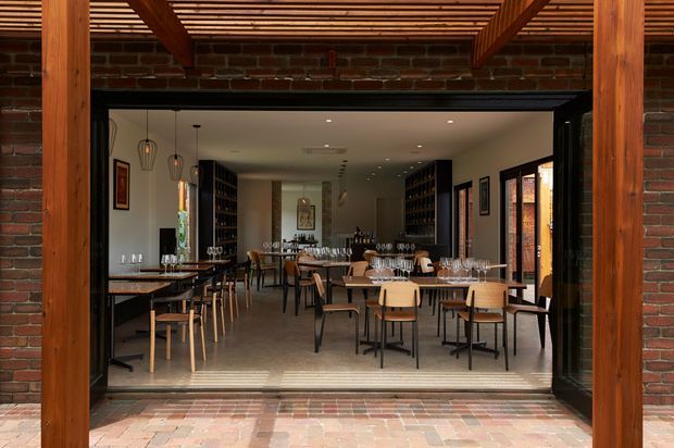 A refined brick-clad hospitality interior viewed from a covered outdoor threshold. Timber posts and a slatted timber ceiling frame large open glazed doors leading into a contemporary dining and wine-bar space. The interior contains multiple timber dining tables and chairs, extensive dark shelving or bottle displays, pendant lights, artwork, and exposed brick detailing. Brick paving continues across the threshold, creating a strong indoor-outdoor connection.