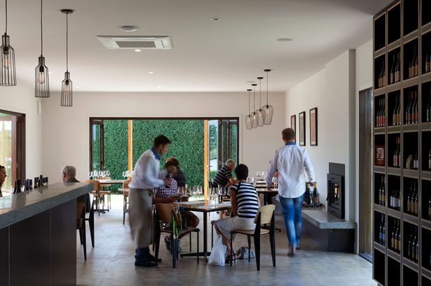 A contemporary dining area or restaurant interior with multiple timber tables and chairs, a long stone-topped counter at left, pendant lighting, polished concrete flooring, and extensive dark built-in shelving displaying bottles. Large glazed doors open toward a planted exterior area, creating a bright, social atmosphere.