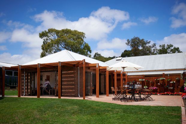 Single-storey pavilion-style building viewed from a landscaped lawn, with timber-framed outdoor structures, dark timber slatted walls, pale corrugated metal roofs, glazed openings, and a brick-paved courtyard furnished with outdoor dining tables and umbrellas. Mature trees and blue sky surround the property.