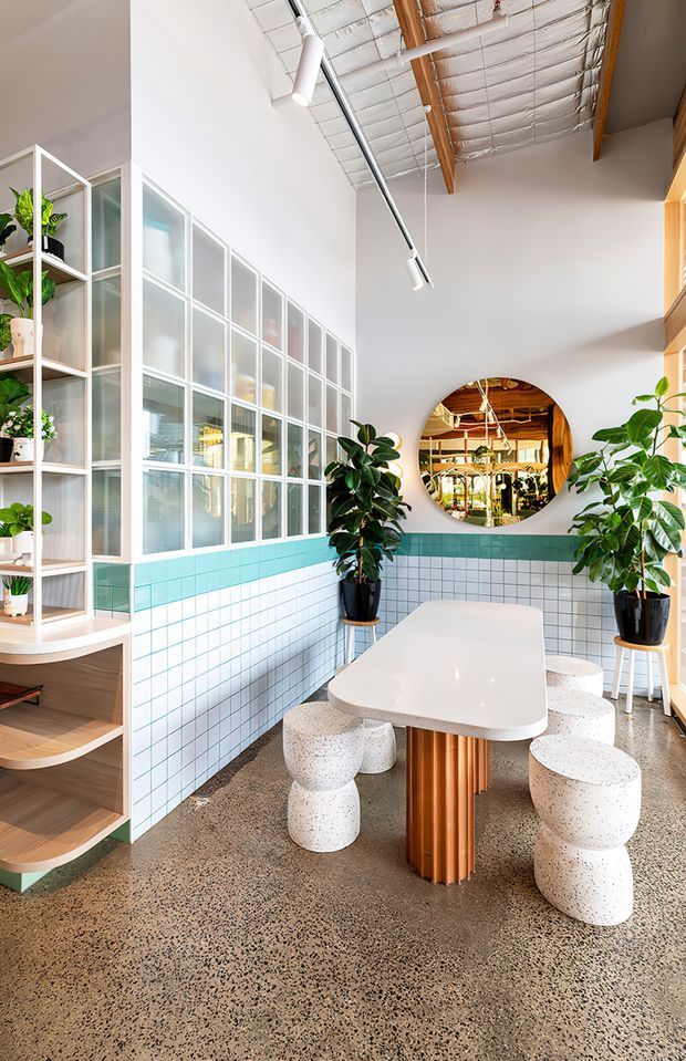 Bright contemporary dining and communal seating area with a long rounded white table, sculptural terrazzo stools, abundant indoor plants, and a large circular mirror. The space features turquoise-and-white tiled walls, extensive translucent framed glazing or partition joinery, exposed timber roof beams with reflective insulation, linear track lighting, and a polished speckled concrete or terrazzo floor.
