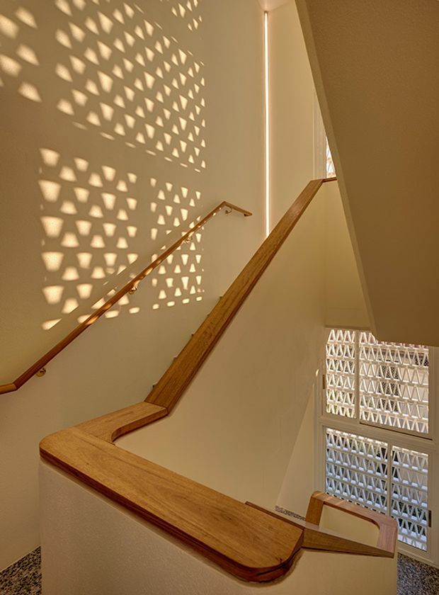 Sunlit interior stairwell with warm timber handrails wrapping around a landing, pale painted walls, and patterned breeze-block screens casting geometric shadows across the stair enclosure. Tall glazing and perforated screens provide strong filtered daylight.