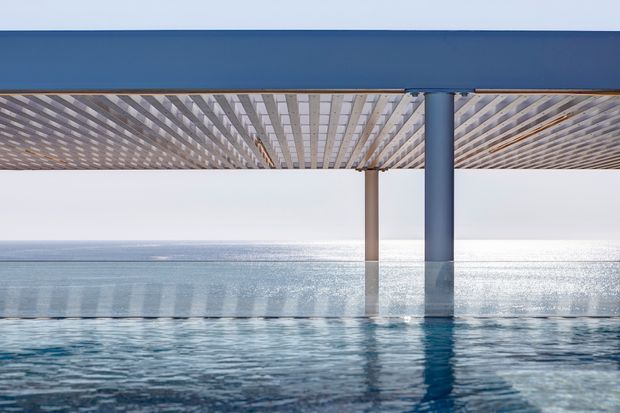 Close-up architectural view of a modern blue-edged pergola or roof structure extending over a swimming pool beside a bright body of water. The underside features a regular slatted or lattice ceiling supported by slender columns, with strong reflections across the pool and a calm coastal horizon beyond.