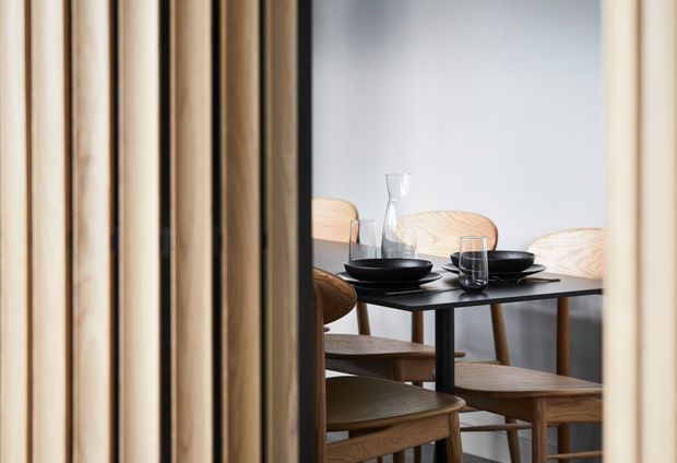 A dining area viewed through a partially open vertical timber screen or slatted partition. A dark rectangular dining table is set with black bowls, plates, glasses and a clear carafe, surrounded by light timber dining chairs. The room has pale walls and a restrained, contemporary natural-material palette.