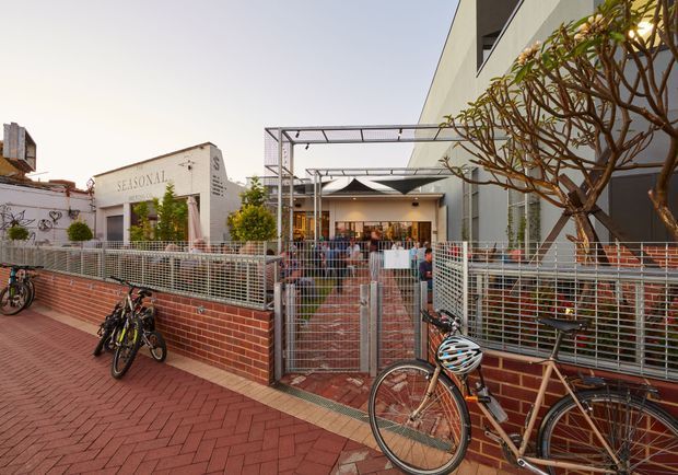 Exterior street-level view of a contemporary mixed-use or commercial venue with a visible entrance courtyard, red brick paving and low brick walls, metal mesh fencing, bicycles parked along the frontage, and a mature frangipani tree. The facade combines a white rendered building with a dark modern wall and a covered outdoor entry structure. A building sign reading “SEASONAL” is visible.
