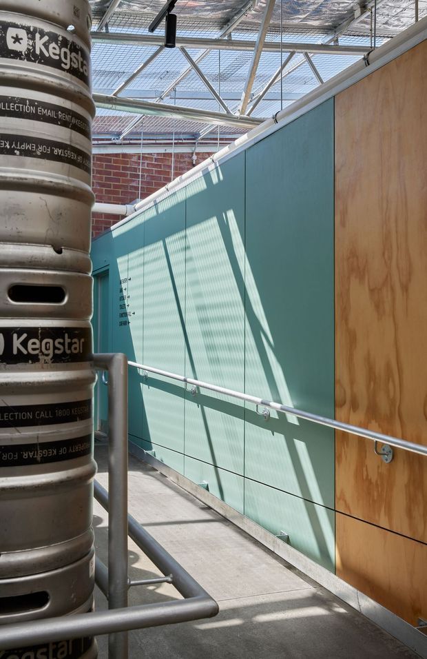 An industrial-style elevated walkway or service corridor enclosed by a glazed and metal-framed roof structure. The corridor has grey concrete flooring, tubular metal guardrails, a teal-green tiled or panelled wall, a plywood-clad wall, and exposed red brick beyond. A stack of beer kegs occupies the left foreground. Strong sunlight filters through the overhead translucent roof, casting linear shadows across the walls and floor.