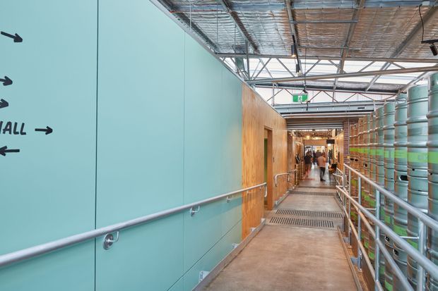 An industrial-style interior walkway or circulation corridor with exposed steel roof framing and services overhead. The left wall is finished in turquoise panels with directional signage and a metal handrail, while the right side contains rows of large cylindrical tanks or industrial vessels behind guardrails. A timber-clad partition and grated drainage flooring lead toward a busier interior area in the distance.