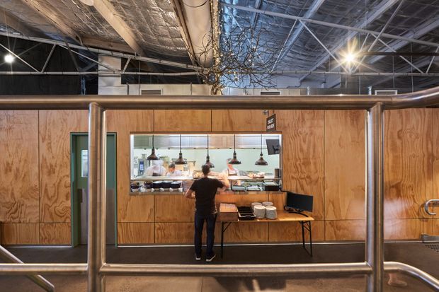 Interior view of an industrial-style commercial food-service space, with a plywood-clad service counter and open kitchen visible behind a large serving window. Stainless-steel pipe railings frame the foreground, while exposed ceiling structure, ductwork, insulation, and pendant lighting reinforce the utilitarian warehouse character.