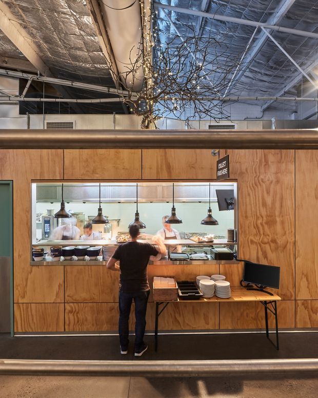 Interior view of a commercial open kitchen or food-service counter. A large serving hatch with glass, multiple pendant heat lamps, stainless-steel preparation surfaces, stacked bowls, a point-of-sale monitor, and staff working behind the counter are set into extensive warm plywood wall panelling. Exposed insulated services and ductwork are visible above, with a decorative branch-and-string-light installation adding a focal feature.