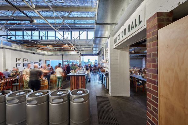 A busy industrial-style commercial dining hall or brewery venue viewed from an open entrance. The space features exposed steel roof framing and services, a high translucent roof, concrete flooring, exposed brickwork, white painted masonry, timber dining furniture, a central bar or service counter, and multiple patrons. A sign identifies the adjoining area as “Function Hall.” Large metal kegs or bins occupy the foreground.