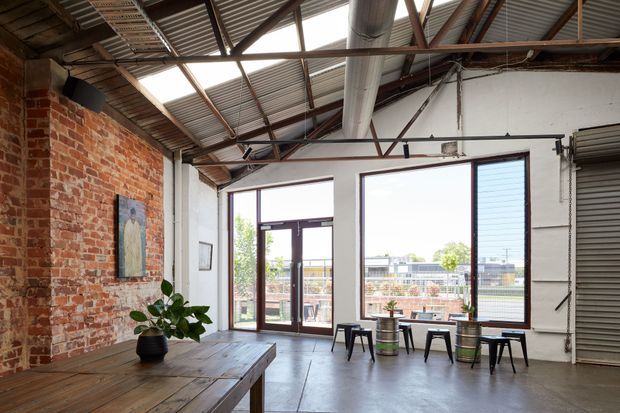 An open industrial-style commercial or studio interior with exposed red brick walls, polished concrete flooring, corrugated metal roofing, exposed steel roof trusses, and large dark-framed windows and glazed doors. A long rustic timber table occupies the foreground, while small stools and barrel-style side tables sit near the glazing. A roller shutter door and visible mechanical ductwork reinforce the warehouse character.