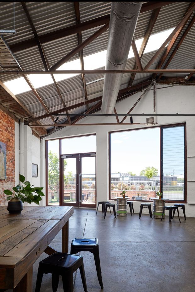 Industrial-style dining and workspace interior with a large timber communal table, black metal stools, polished concrete flooring, exposed brick and plaster walls, and expansive steel-framed glazing opening to an outdoor terrace. The high corrugated metal roof features exposed dark timber and steel trusses, skylights, and a prominent exposed ventilation duct.