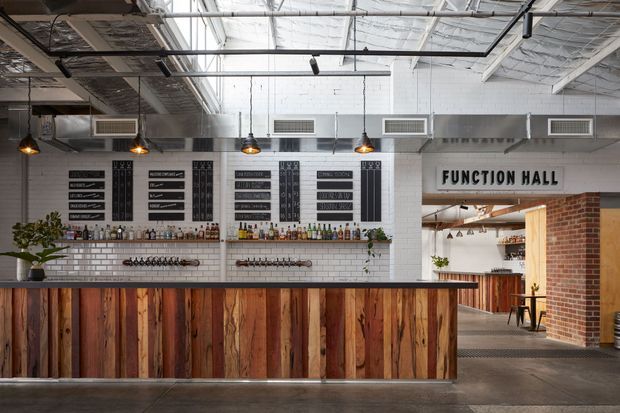 A large industrial-style function hall bar with a long timber-fronted counter, exposed services and ductwork, white painted brick walls, black track lighting, hanging pendant lights, and extensive bottle and glass storage. A sign reading “FUNCTION HALL” opens toward an adjoining bar area with exposed brickwork and timber finishes.