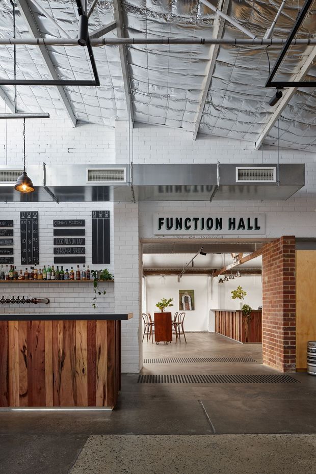 Interior view of a converted industrial-style function hall and bar. The space features exposed roof framing with reflective insulation, suspended services and track lighting, white painted brick walls, stainless-steel ventilation ductwork, a timber-faced bar, polished concrete flooring, and a brick-framed opening leading to a dining and service area. Chalkboard menus and beverage shelving reinforce the hospitality venue setting.