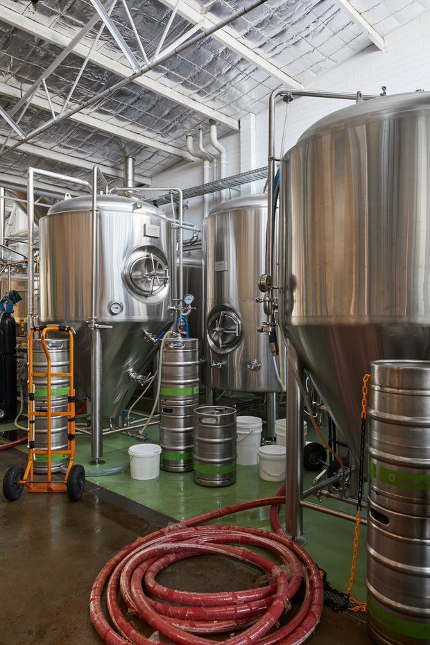 Industrial brewery interior filled with large stainless-steel fermentation and brewing tanks, interconnected metal piping, kegs, hoses, and utility equipment. The space has a high exposed industrial ceiling with reflective insulation, white painted masonry walls, and a green-painted working floor. Bright functional lighting and dense brewing equipment dominate the scene.