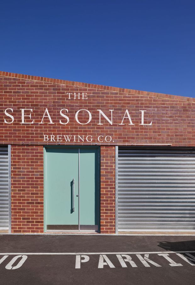 Exterior street-level view of a brewery building with a red brick facade, large painted signage reading “The Seasonal Brewing Co.”, pale blue-green glazed entrance doors, and a silver-grey roller shutter. Angled roofline, asphalt forecourt, and painted parking markings are visible under a clear blue sky.