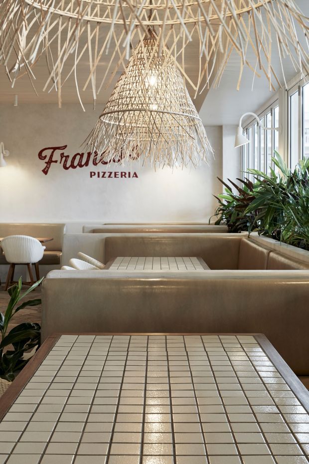 A bright contemporary pizzeria interior with built-in tan upholstered booths, tiled dining tables, white sculptural chairs, abundant indoor plants, and large windows along one side. The focal feature is an oversized woven, organic pendant installation suspended from the ceiling, complemented by a smaller woven pendant over the rear seating. A red script pizzeria sign is mounted on a pale plaster wall.