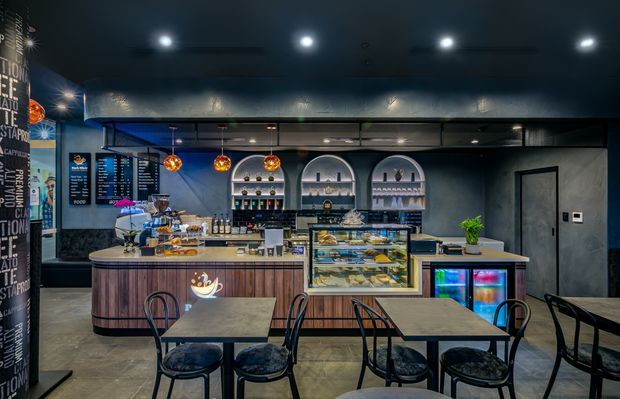 A modern café interior with a compact dining area in the foreground and a prominent service counter behind it. The space features dark grey walls and ceiling, polished concrete-style flooring, timber-fronted curved counter cabinetry, black metal-framed furniture, illuminated pastry and beverage displays, arched wall shelving, pendant lights, and menu boards.
