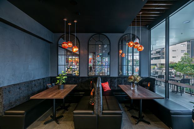 A dark, refined commercial dining interior with booth seating, timber tables, black upholstered banquettes, arched steel-framed windows, pendant globe lighting, and exposed timber ceiling battens. Large glazing opens visually to an outdoor terrace and urban surroundings.
