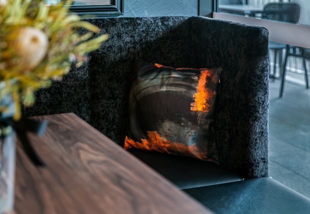 Close-up interior detail of a dark upholstered booth or banquette beside a richly grained dark timber table. A black, brown, and vivid orange patterned cushion provides a strong accent, with a blurred floral arrangement in the foreground and glimpses of dining furniture in the background. The mood is dark, refined, and contemporary.