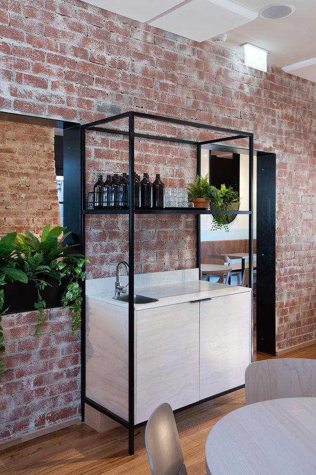 A compact open kitchenette or bar area framed in black metal shelving, with pale wood cabinetry, a light benchtop, integrated sink and tap, and dark bottles and glassware on the open shelves. The space has exposed red brick walls with white mortar, timber flooring, black-framed openings, potted greenery, and a partial round dining table with chairs in the foreground. A dining area is visible beyond the opening.