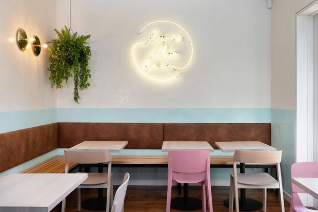 A compact, brightly styled café or commercial dining space with built-in booth seating along the wall, small square dining tables, and mixed pastel chairs. The walls feature a two-tone white and pale aqua paint scheme with a dark brown upholstered or leather-look horizontal wall band. A hanging fern, round wall lights, and a glowing neon-style sign add decorative accents. Dark timber flooring and large glazing at the right complete the contemporary interior.