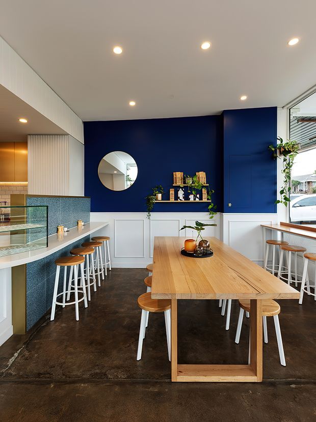 Bright, contemporary dining and kitchen interior with a long light-oak communal dining table and matching stools, a deep navy blue paneled feature wall, white wainscoting and cabinetry, a blue-speckled tiled island, dark polished concrete flooring, and recessed ceiling lights. A round mirror, open shelving, indoor plants, and large windows add detail and natural light.